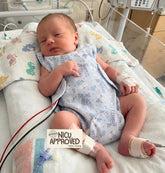 Premature baby boy in hospital crib wearing Blue Bowy print NICU romper. Patent-pending design shows easy access for monitoring leads and wires. Duper DuDu brand