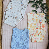 Flatlay of the NICU Essentials Trio bundle by Duper DuDu. Three adaptive baby onesies laid on a soft pink muslin cloth in a woven basket. The set includes three patterns: a cream 'Cutie buds' print, a blue 'Bowy' print, and a yellow 'Bakery Dreams' print. All feature GOTS organic cotton fabric, soft scalloped trims, and the nurse-designed Huggle Pad side-opening for easy IV access.
