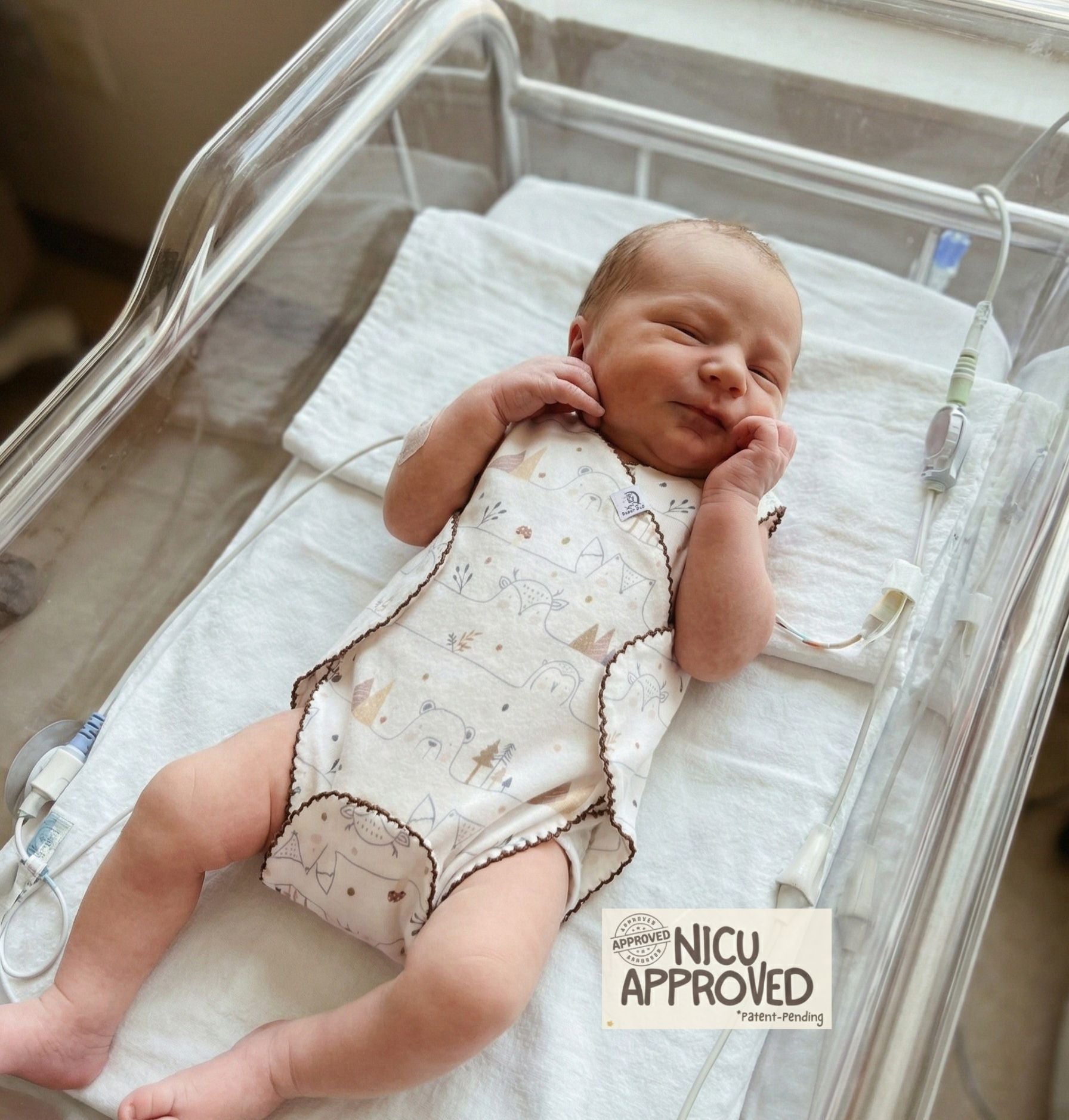 Premature baby in NICU incubator wearing adaptive organic cotton romper. Design allows easy access for medical monitoring wires. NICU Approved