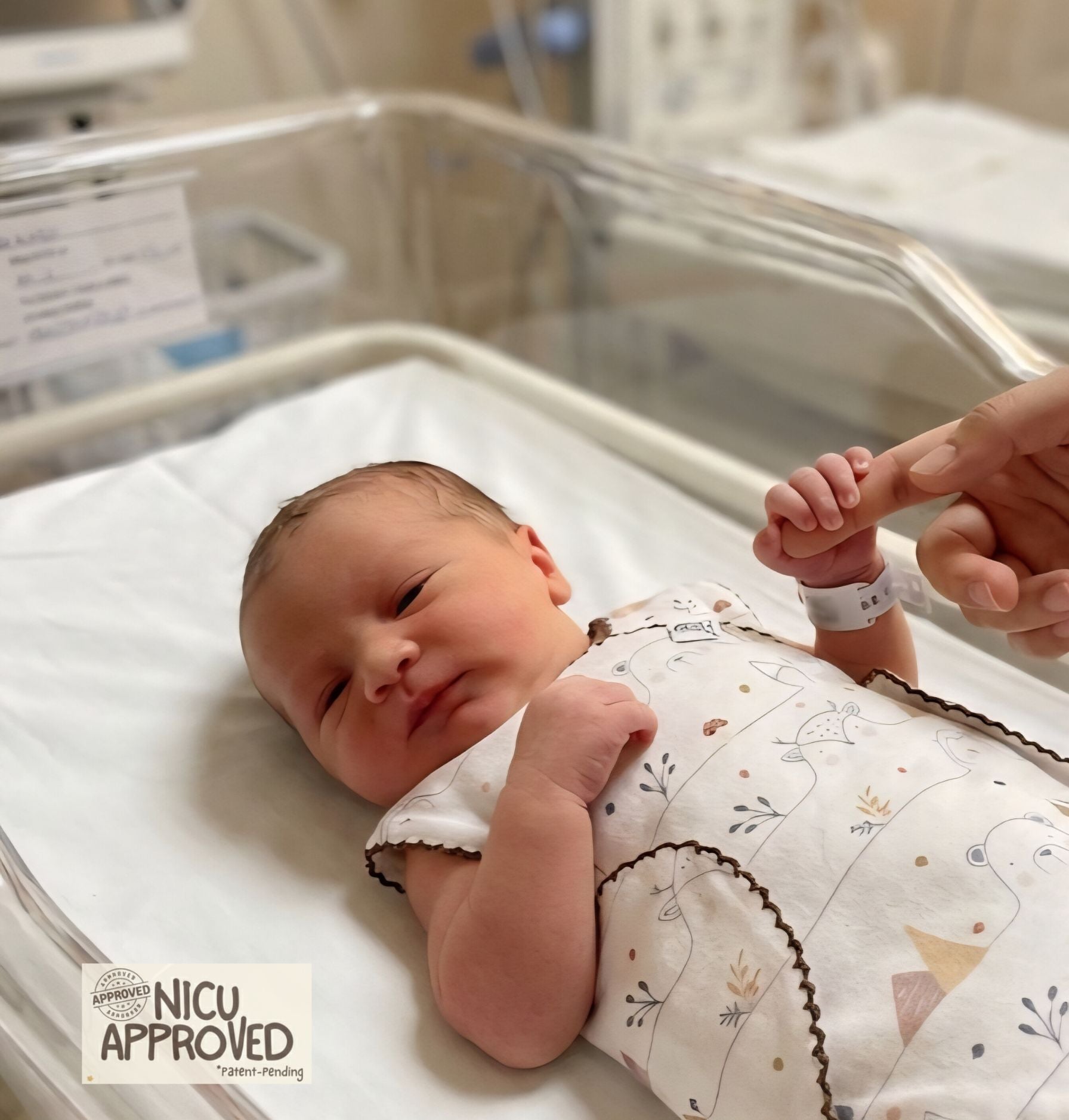 Newborn baby in a hospital crib with a hand holding mom's hand, NICU Approved label visible. Wearing organic preemie onesie brand Duper DuDu.