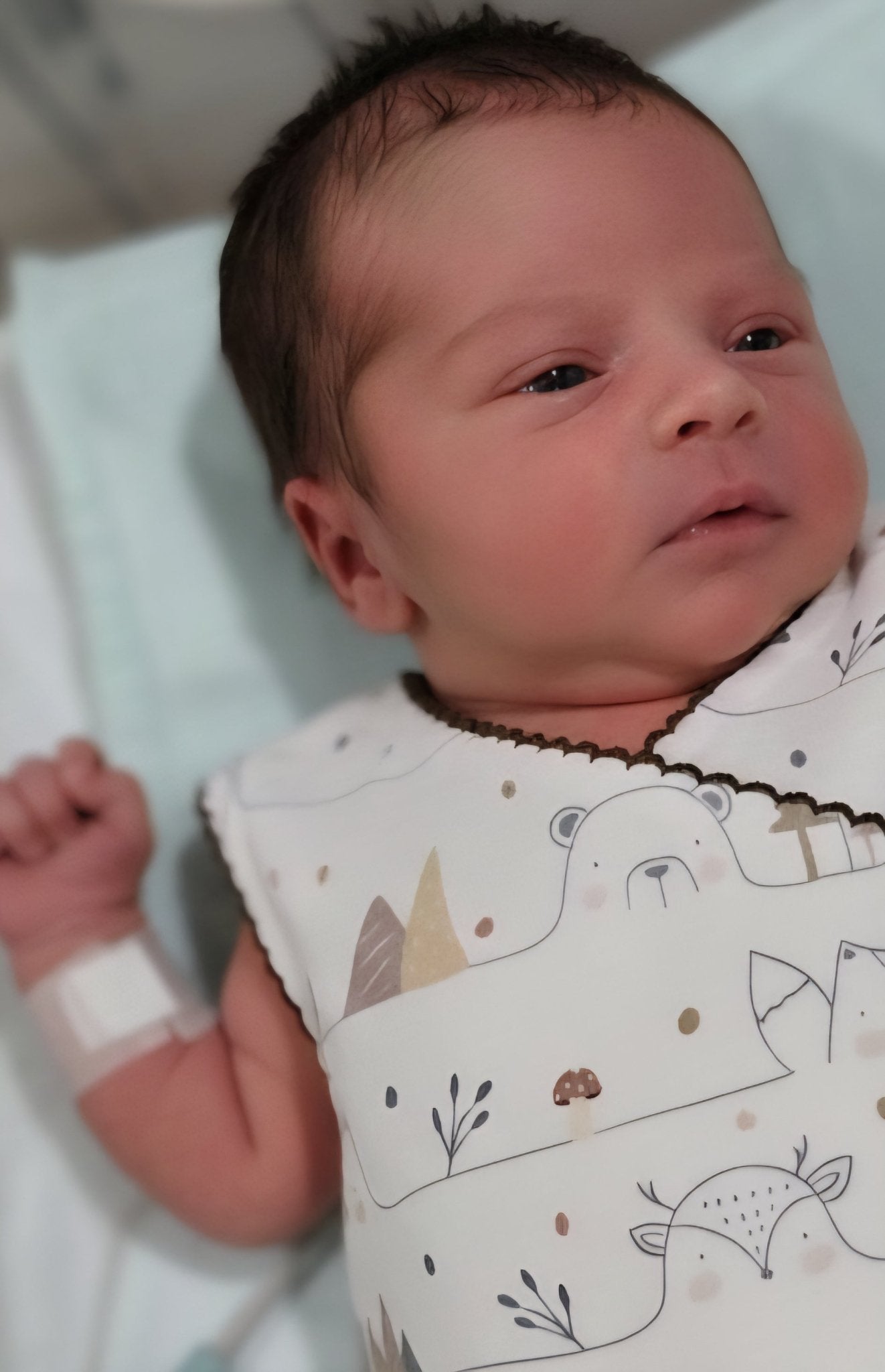 Close-up of premature baby face looking comfortable in organic cotton NICU romper. Hospital wristband visible on tiny arm. Soft breathable fabric with bear pattern. Brand Duper DuDu