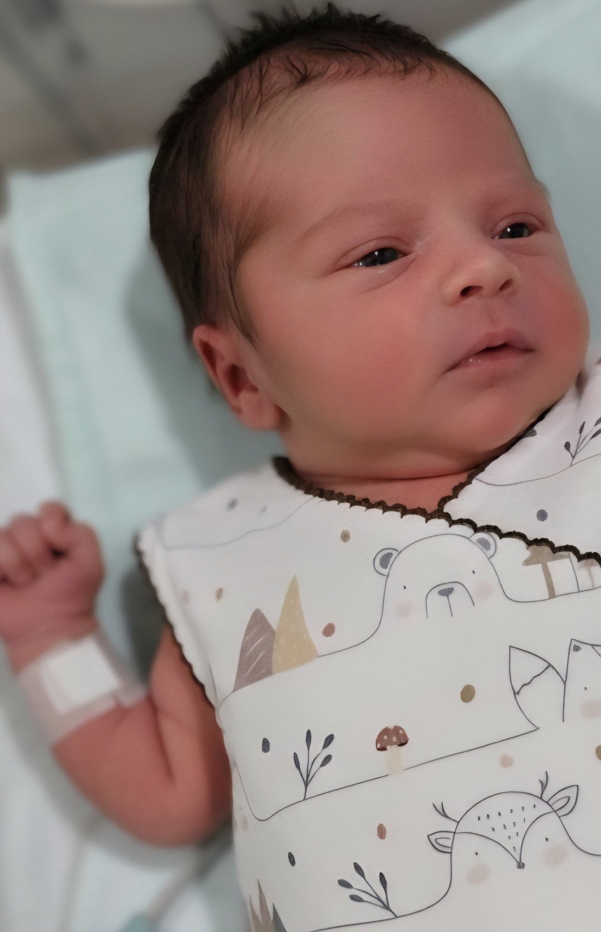 Close-up of premature baby face looking comfortable in organic cotton NICU romper. Hospital wristband visible on tiny arm. Soft breathable fabric with bear pattern. Brand Duper DuDu