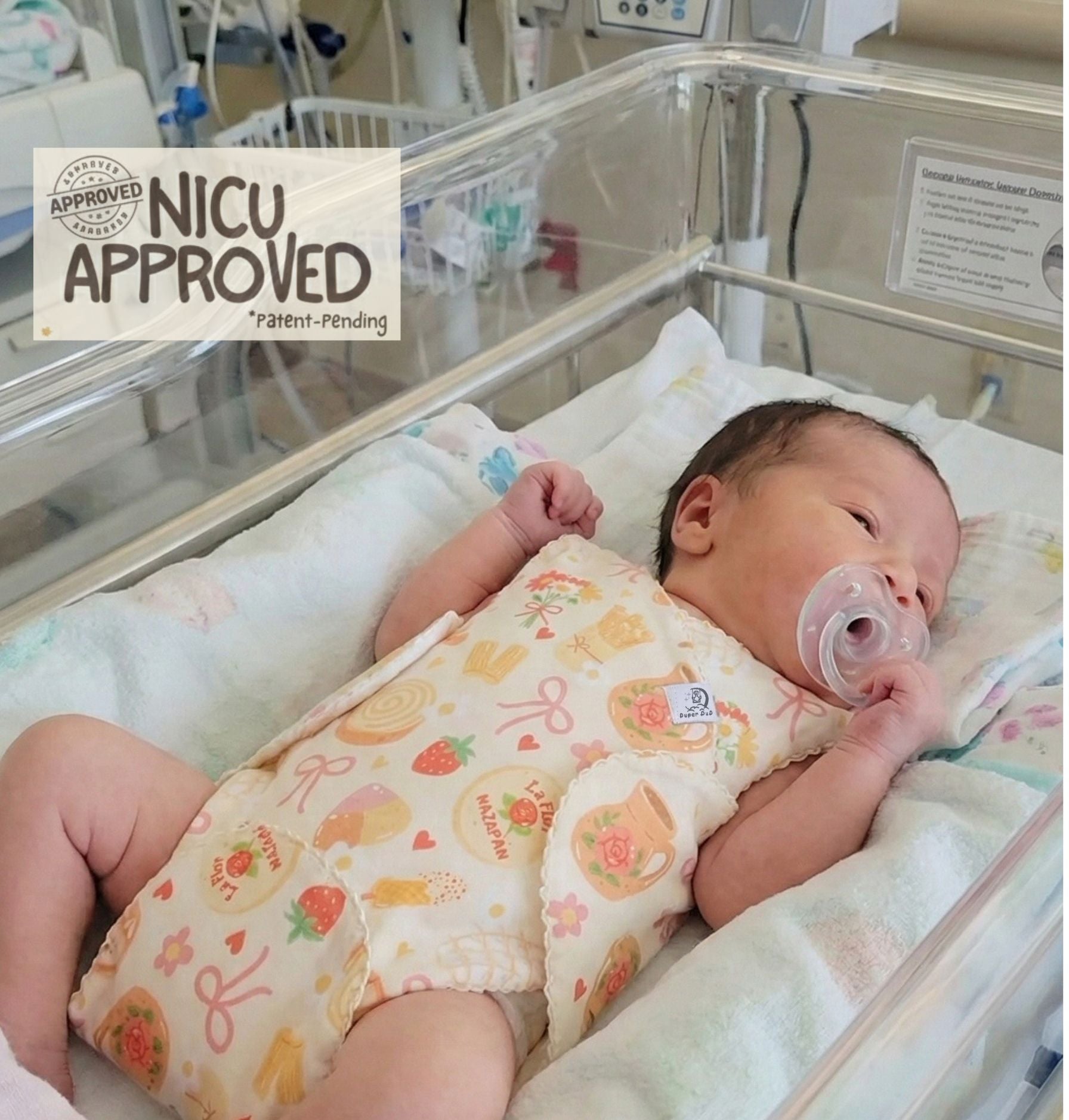 Newborn baby resting in hospital crib wearing patent-pending NICU romper brand Duper DuDu (Bakery Dreams print). Easy-access design for nappy changes without disturbing baby.